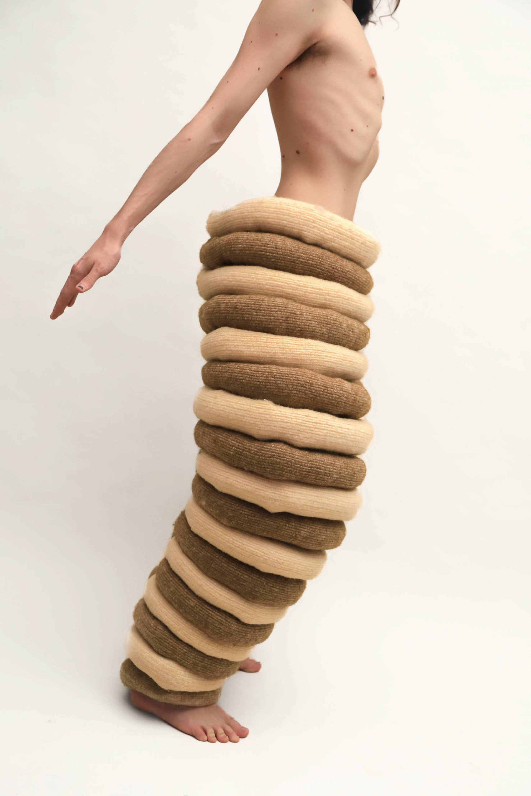 A person in profile wearing sculptural pants composed of alternating beige and brown padded rings, extending from waist to ankles, standing barefoot against a plain background.