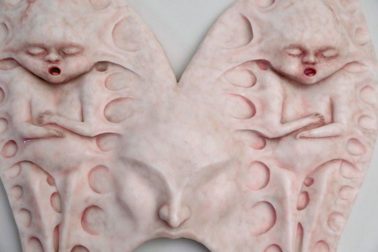 Silicone headpiece shaped like two winged forms joined at the center, each containing a small sculpted baby figure in relief. The piece covers the wearer’s head and face, merging organic and mechanical forms.