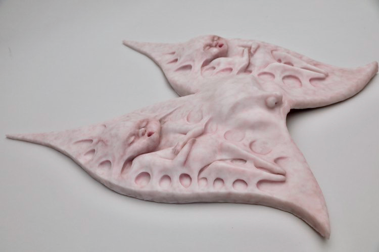 Silicone headpiece shaped like two winged forms joined at the center, each containing a small sculpted baby figure in relief. The piece covers the wearer’s head and face, merging organic and mechanical forms.