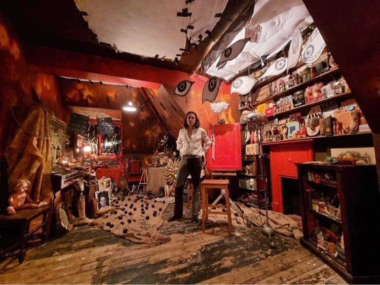 A dim attic studio cluttered with dolls, paintings, masks, and scattered art materials under warm red and yellow light.