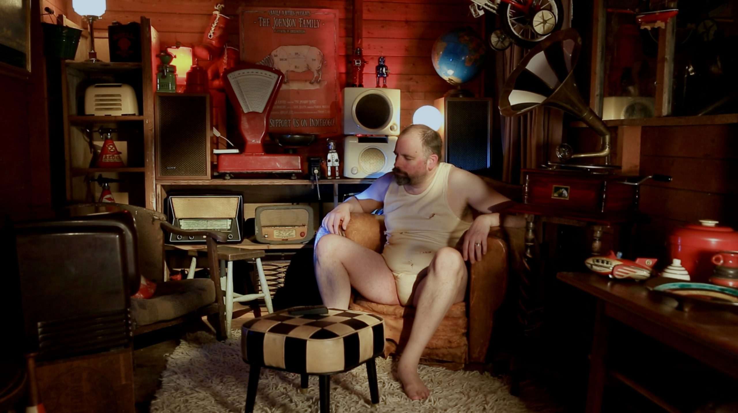 A beer bellied man in an undershirt sits in a cluttered retro room filled with vintage radios, scales, and lamps.