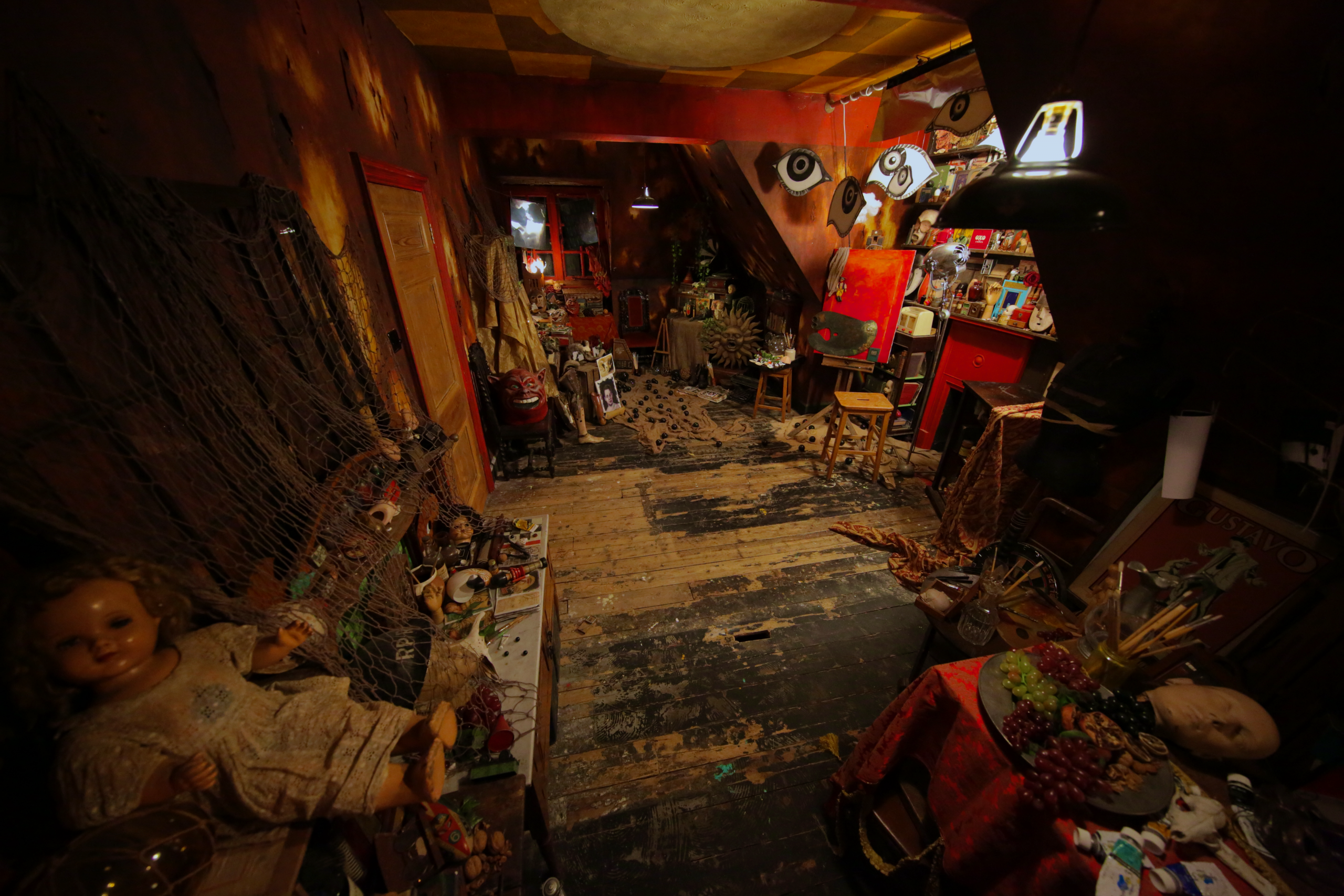 A dim attic studio cluttered with dolls, paintings, masks, and scattered art materials under warm red and yellow light.