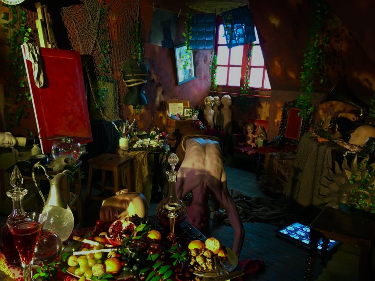 A dimly lit, surreal artist’s studio filled with mannequins, masks, plants, and a figure bending forward among scattered objects and fruit.