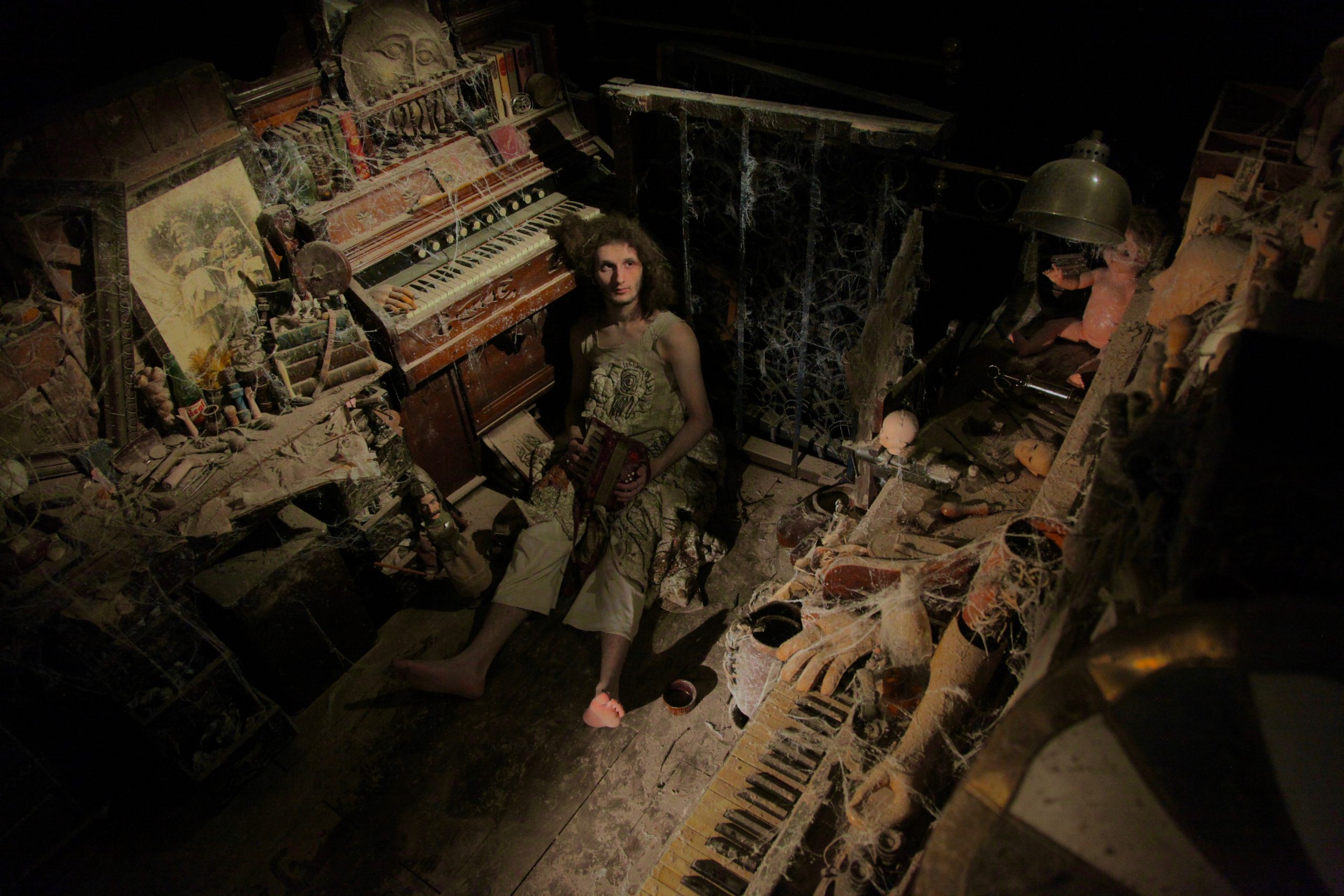 A barefoot figure sits among cobweb-covered instruments and dolls in a dark, decaying room.