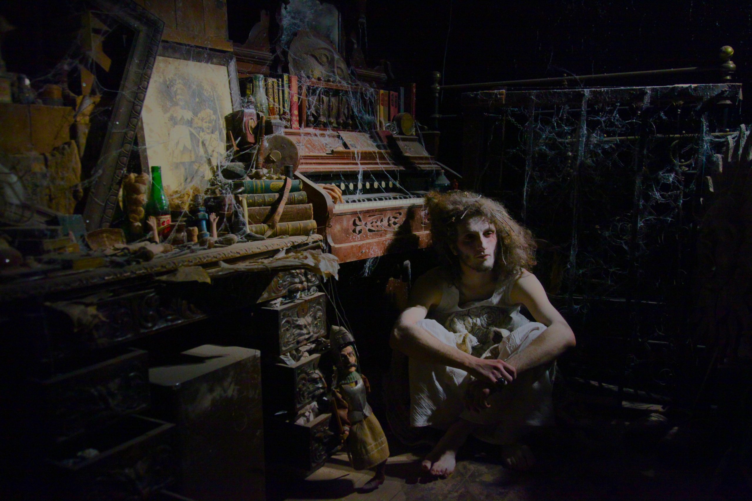 A person with disheveled hair sits barefoot on the floor beside a dusty organ in a dark, cluttered room filled with cobwebs and old objects.