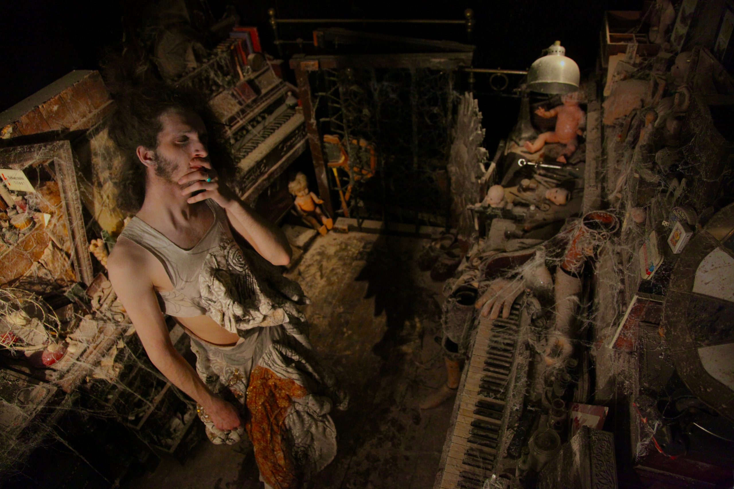 A person stands in a dim, dust-covered room filled with cobwebbed instruments, dolls, and debris.