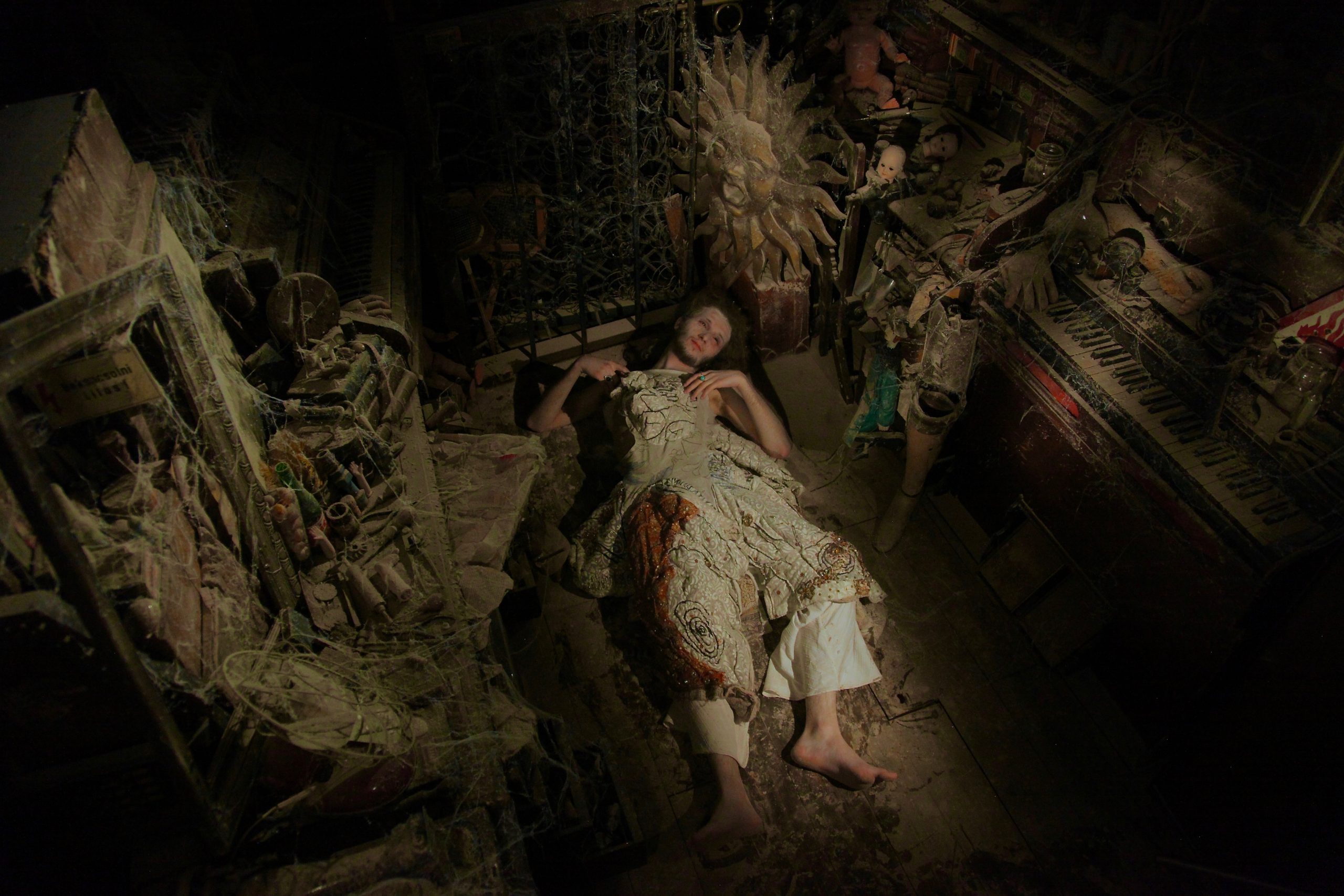 A person lies on the floor of a dark, dusty room filled with cobwebs, surrounded by decaying musical instruments and doll parts.