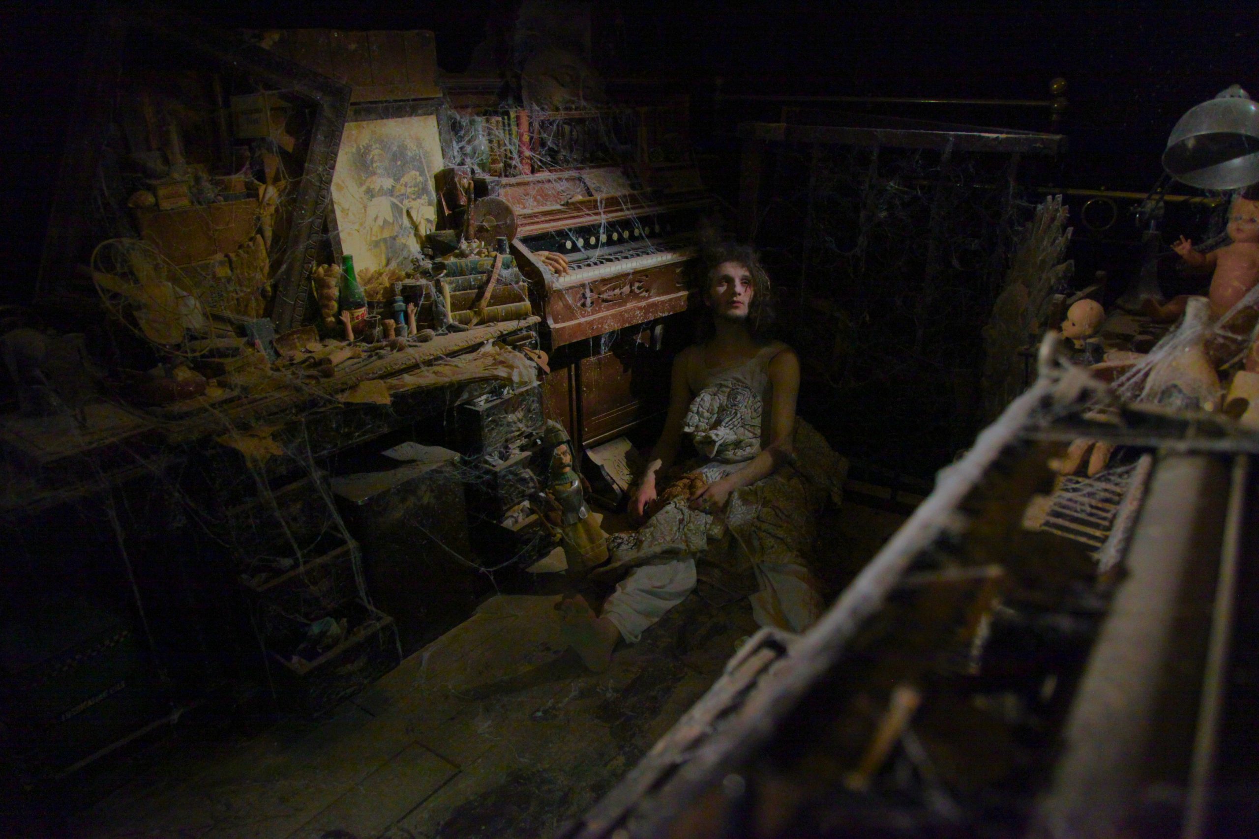 A person sits on the floor of a dark, dust-covered room, looking upward among cobwebbed instruments and decaying objects.
