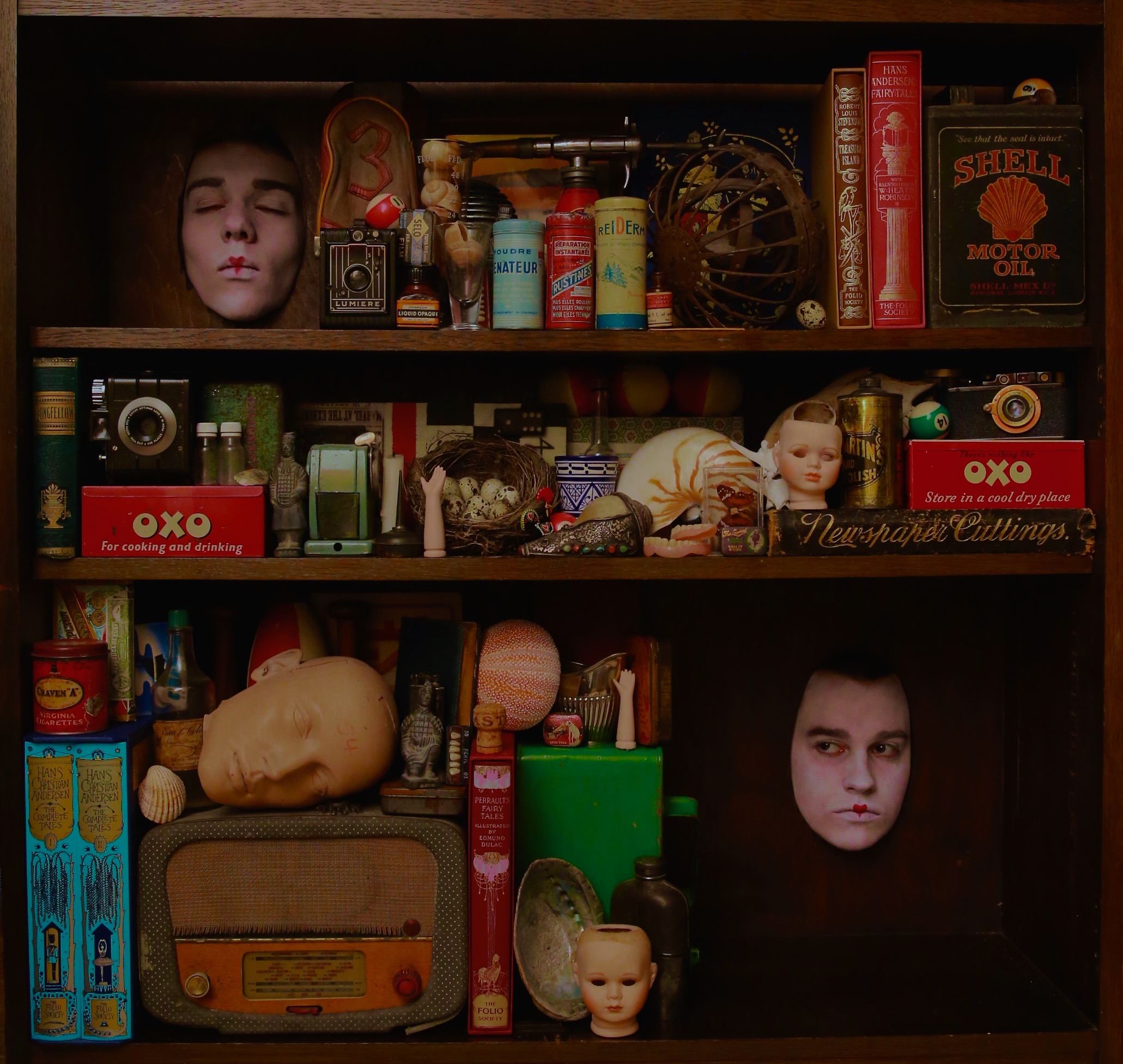 A wooden cabinet filled with vintage objects, dolls’ heads, cameras, and two human faces emerging among the items.