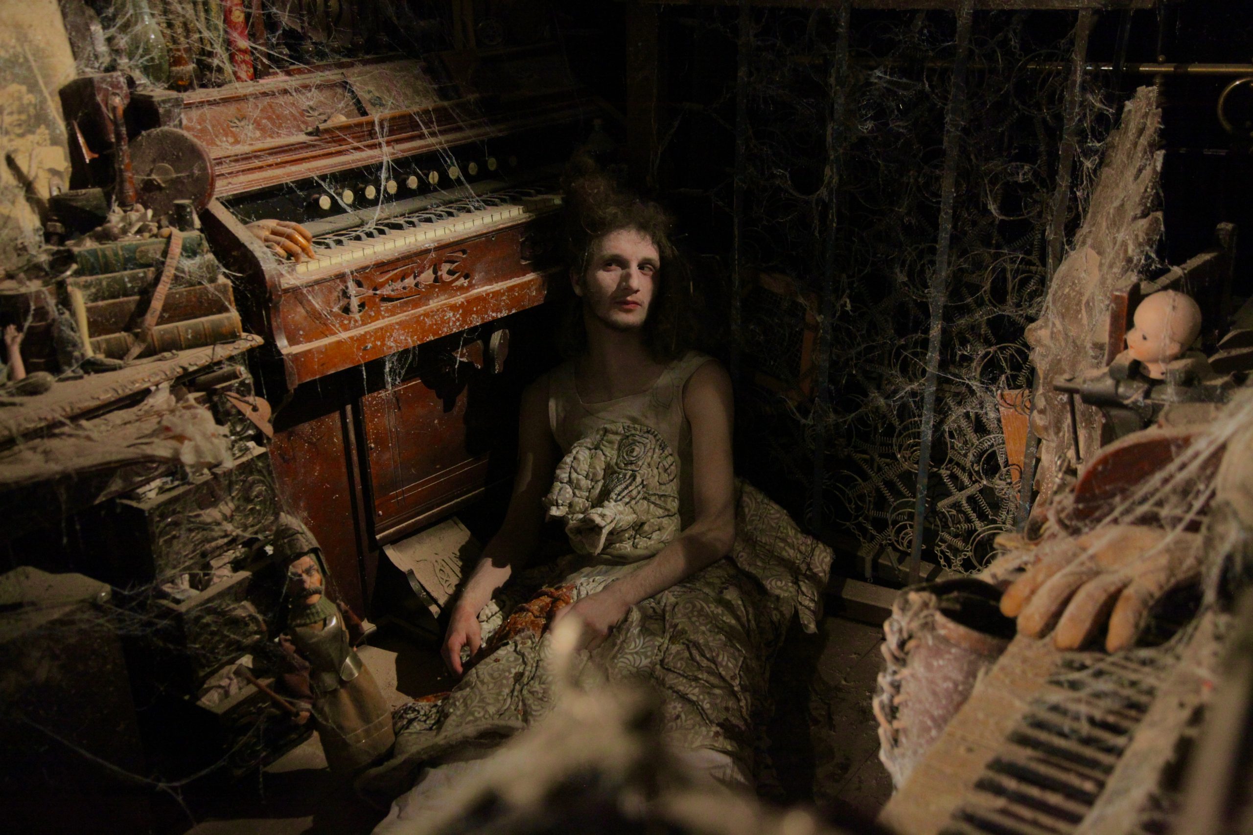 A person sits on the floor beside a dusty organ in a dark, cobweb-covered room, looking slightly upward through the gloom.