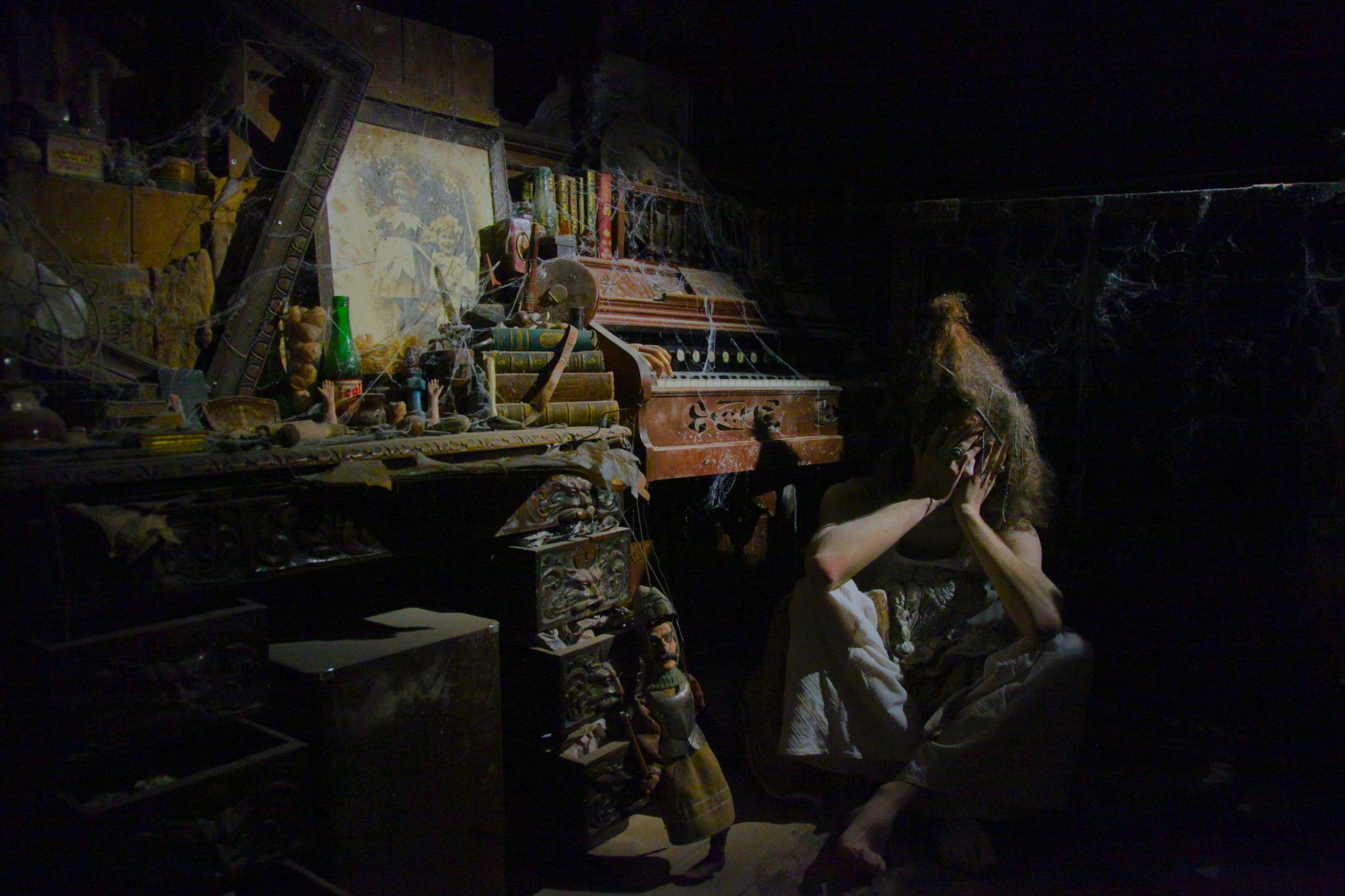 A person sits on the floor of a dim, cluttered room, covering their face with their hands beside a dust-covered organ and cobwebbed objects.