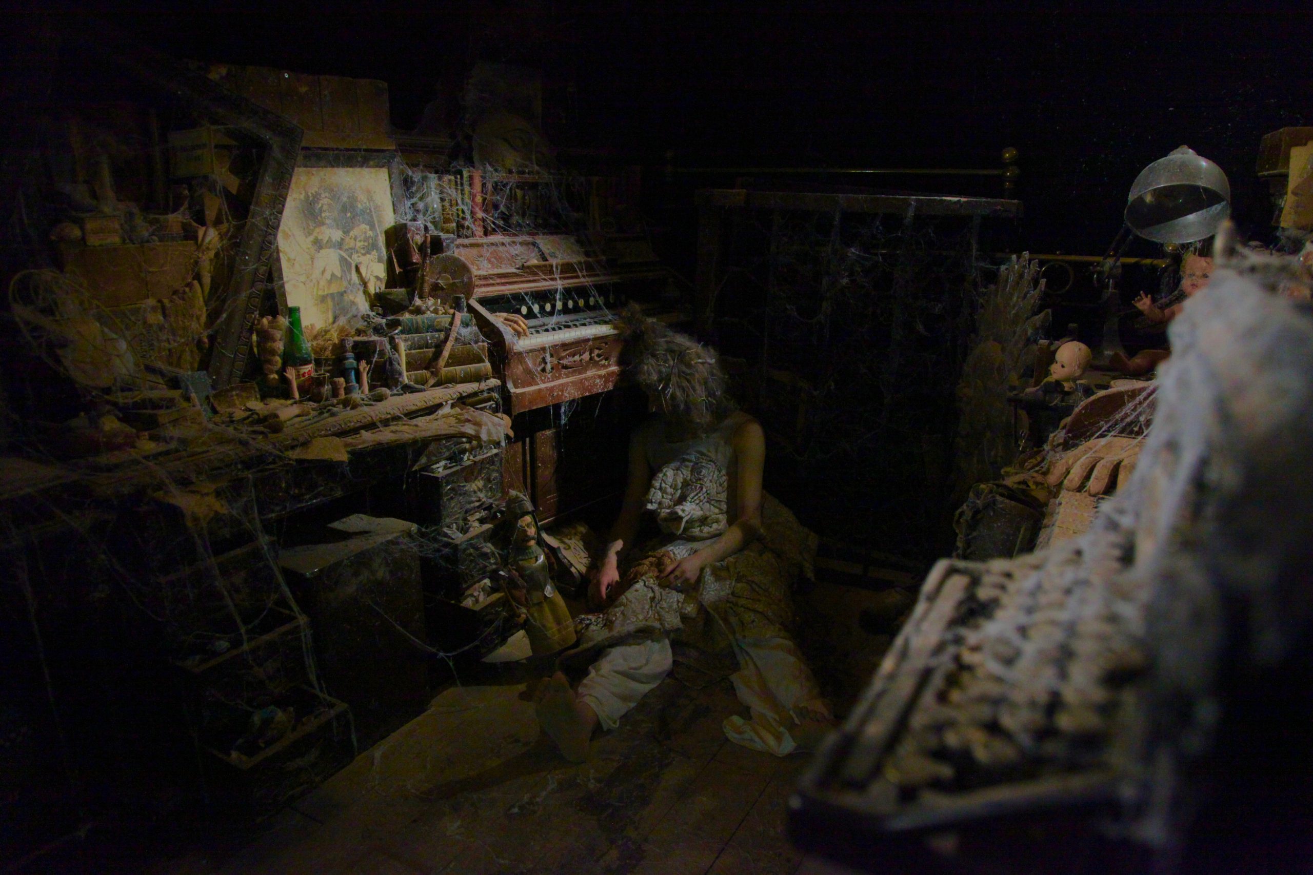 A person sits slumped in a dim, dust-covered room filled with cobwebbed instruments and decaying objects.
