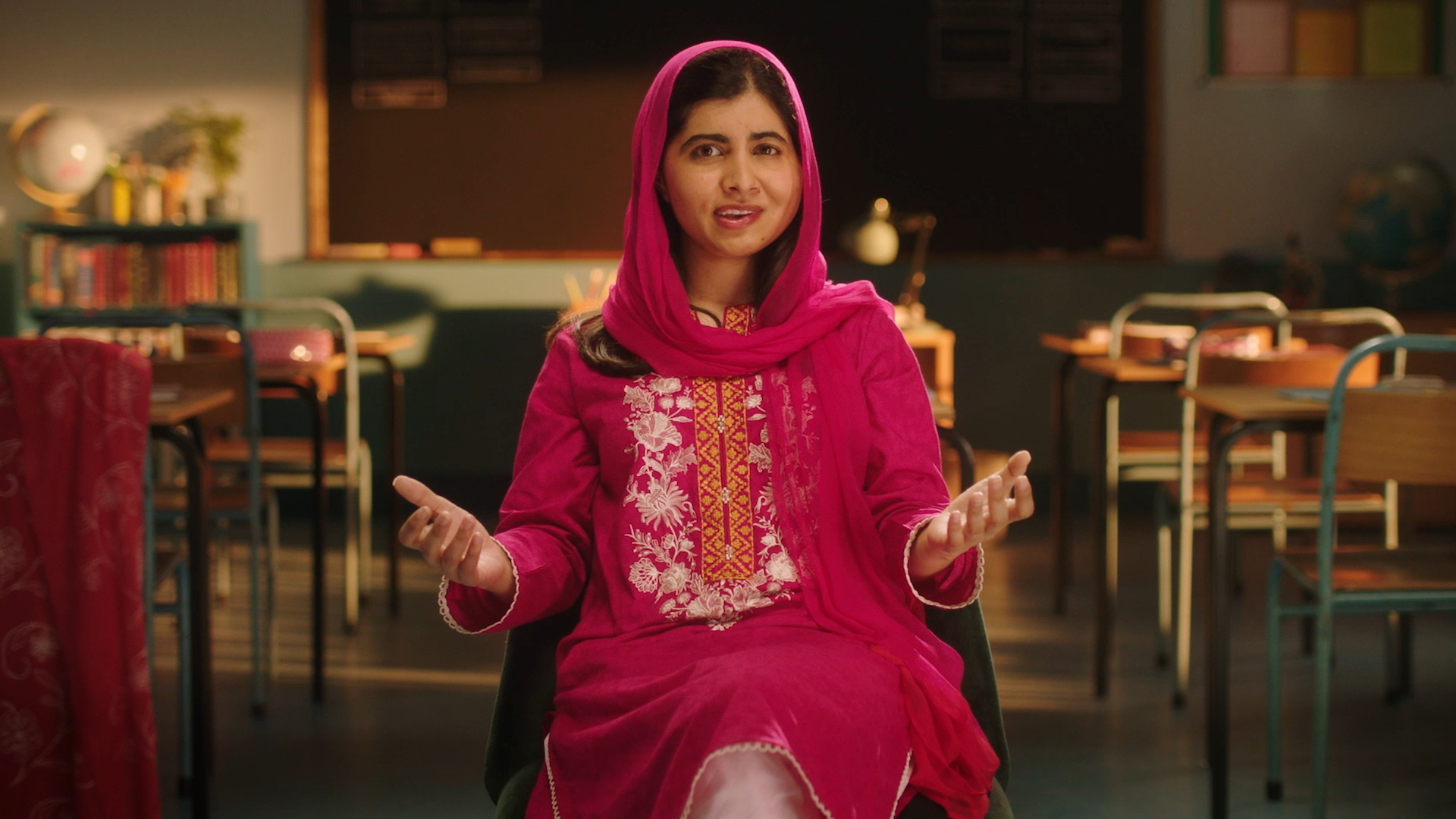 Malala Yousafzai sits in a recreated classroom set, wearing a bright pink outfit with embroidered details, speaking directly to the camera.