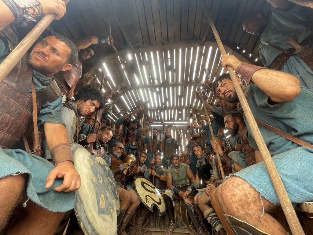 A group of men dressed as ancient warriors sit tightly packed inside a wooden structure, spears and shields raised around them. Light filters through slats in the ceiling, casting striped beams across their worn faces and armor. Their expressions are tense and focused, evoking the anticipation of battle in a confined, almost sacred space.