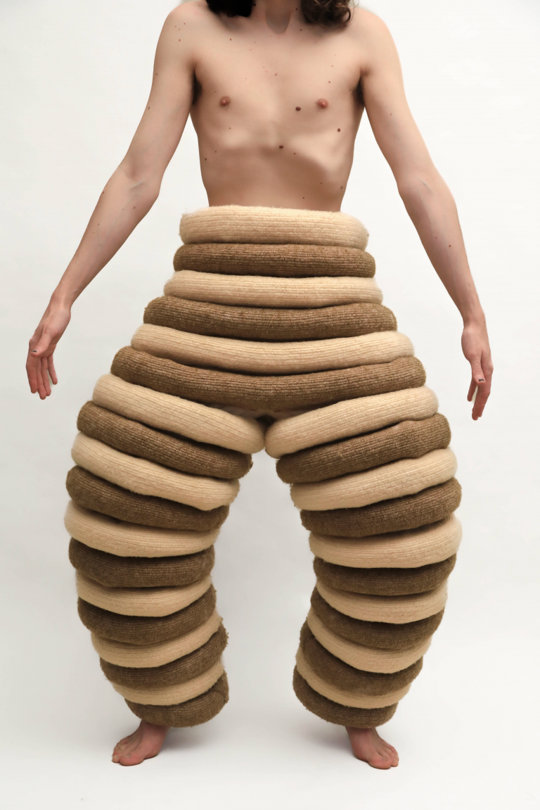 A person stands against a plain background wearing sculptural, ringed pants made of alternating bands of beige and brown knitted fabric, resembling stacked coils.