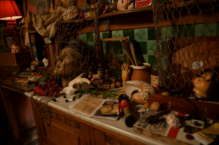 A dimly lit, cluttered artist’s workspace filled with eclectic objects. On the marble-topped wooden counter are sketchbooks open to portrait drawings, small bottles, a human skull replica, a ceramic pot holding paintbrushes, a wooden hand model, walnuts, a nautilus shell, and a vintage toy. A sculpted head draped in a fishing net and artificial grapes rests beside the skull. Behind the counter, green tiled walls and wooden shelves display more objects—dolls, jars, books, and a red lamp casting warm light over the scene. The atmosphere feels nostalgic and mysterious, evoking the workspace of a collector or eccentric artist.
