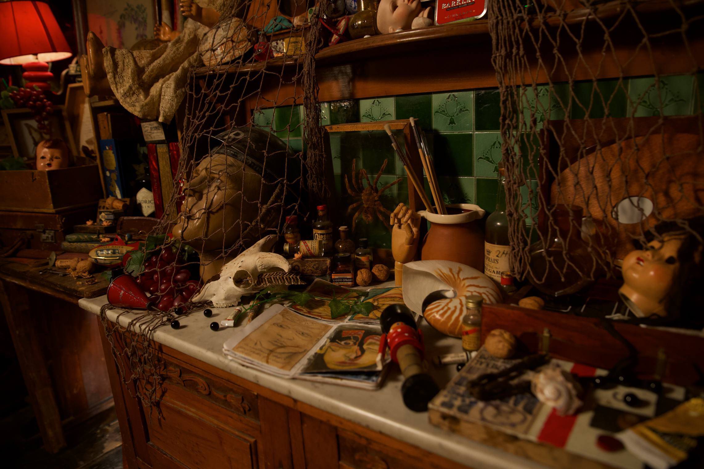 A dimly lit, cluttered artist’s workspace filled with eclectic objects. On the marble-topped wooden counter are sketchbooks open to portrait drawings, small bottles, a human skull replica, a ceramic pot holding paintbrushes, a wooden hand model, walnuts, a nautilus shell, and a vintage toy. A sculpted head draped in a fishing net and artificial grapes rests beside the skull. Behind the counter, green tiled walls and wooden shelves display more objects—dolls, jars, books, and a red lamp casting warm light over the scene. The atmosphere feels nostalgic and mysterious, evoking the workspace of a collector or eccentric artist.