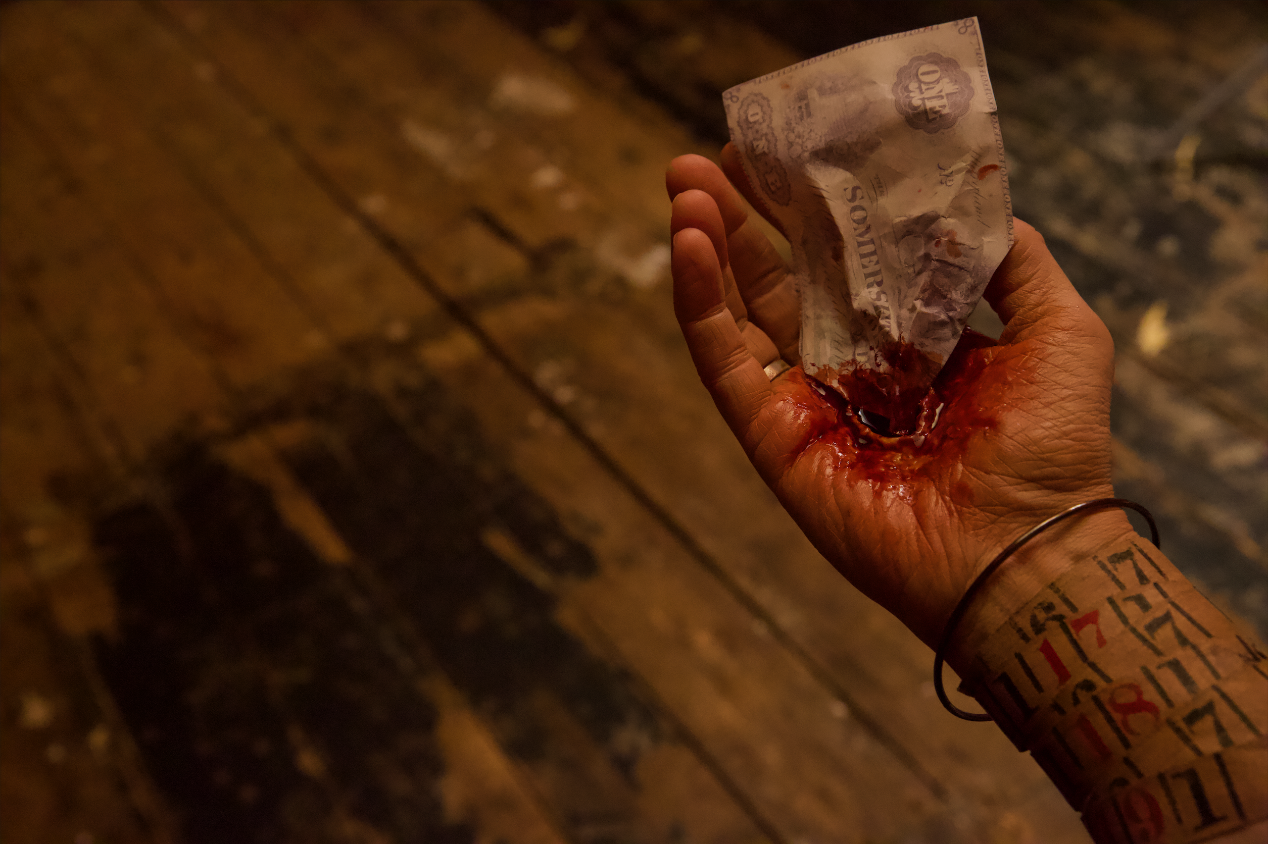A close-up of a person’s hand against a worn wooden floor. The palm appears to have a realistic-looking special-effects wound with red coloring resembling blood, holding a crumpled paper currency note partially inserted into it. The person wears a silver bracelet and has bold black and red lettering tattooed on their wrist and forearm. The lighting is dim and dramatic, emphasizing the contrast between the skin, the red coloring, and the rough wooden background.